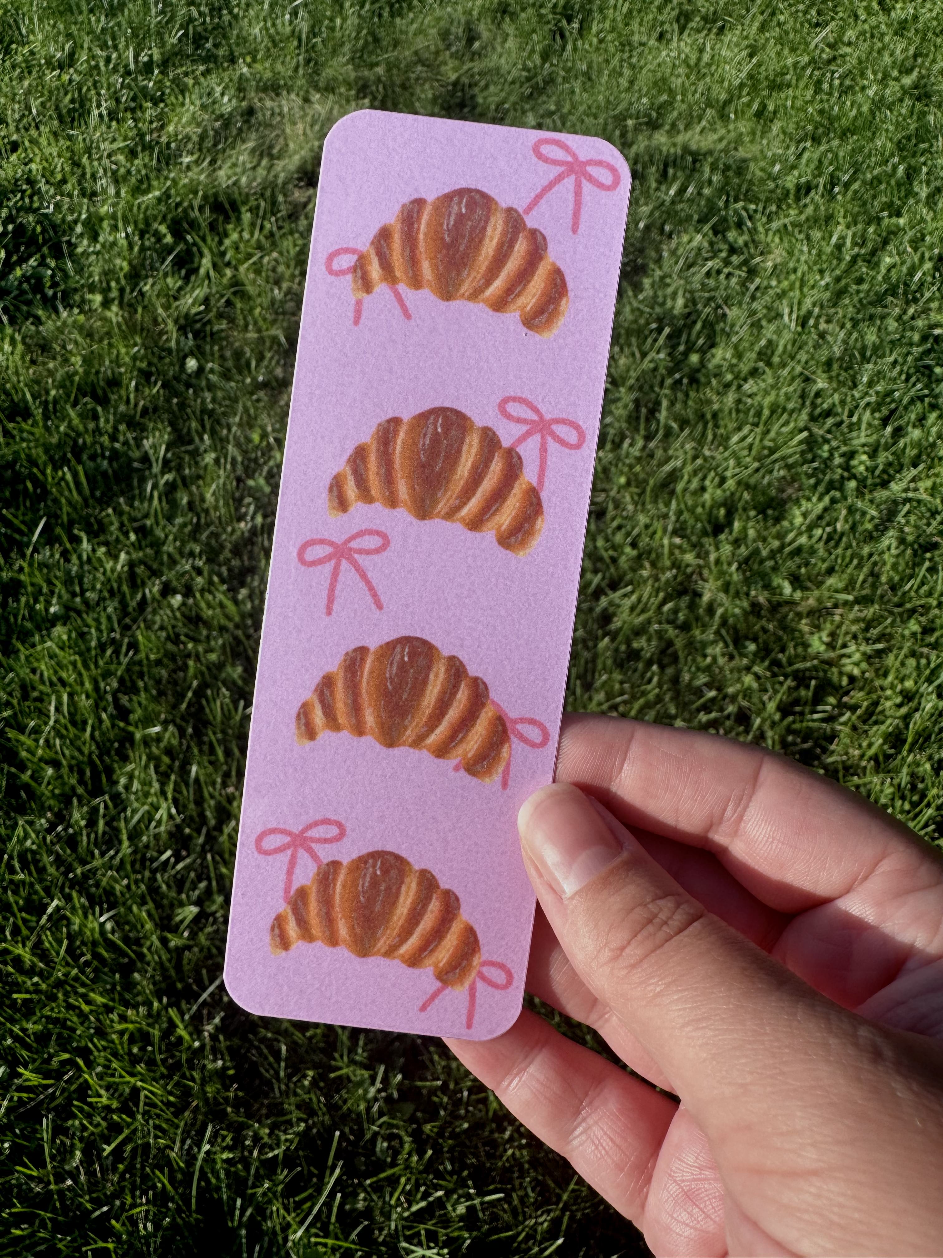 Croissant and bows oil pastel bookmark on lavender background held against green grass