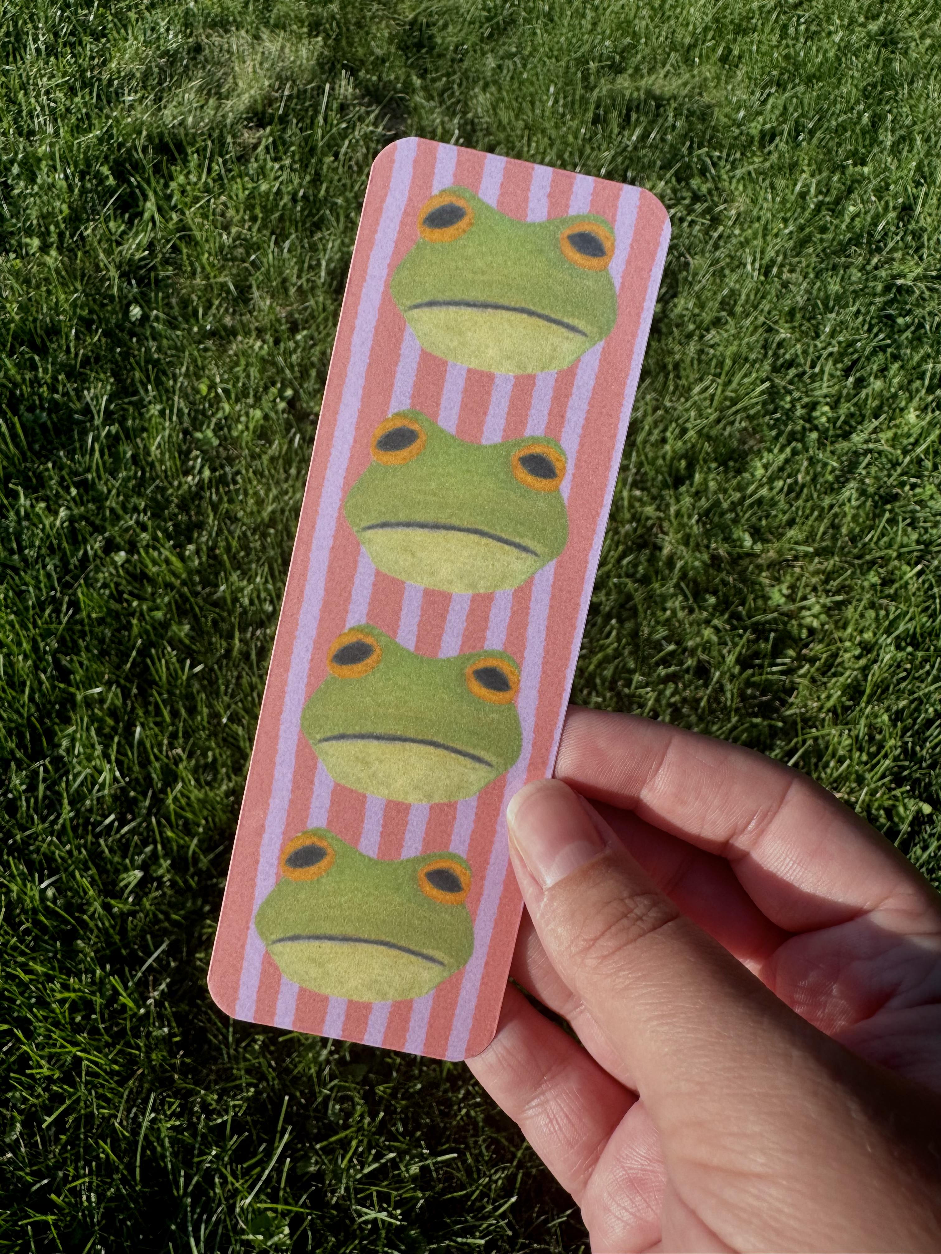 Frog bookmark with four round green frogs with golden eyes on pink and lavender stripe background